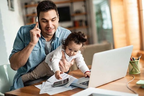 Papá usando la computadora portátil mientras habla en el móvil y sostiene a un niño curioso