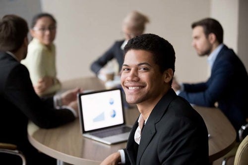 hombre sonriendo con la computadora portátil en la mesa con otras personas con atuendos de negocios